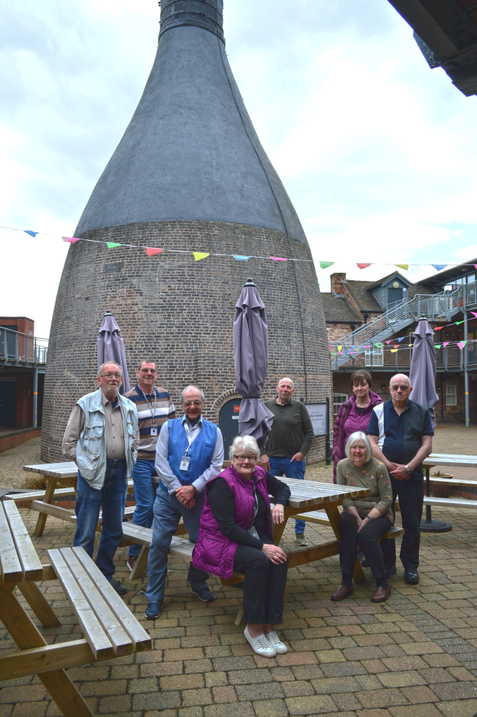 Group of eight older adults posing around wooden picnic benches in an outdoor courtyard, in front of a large bottle-shaped brick kiln. Colourful bunting is strung across the space, with closed patio umbrellas and surrounding red-brick buildings visible in the background under a cloudy sky.